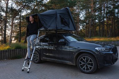 Katlanabilir çatı çadırı olan koyu renkli bir SUV bir ormanda asfalt bir yüzeye park edilmiştir. Bir adam sıcak doğal ışık altında çadıra merdiven çıkıyor..