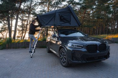 Çadırı katlanabilir modern bir SUV ormanda park halinde. Sıcak ışık uzun ağaçların arasından süzülürken bir insan çadıra merdiven tırmanır..