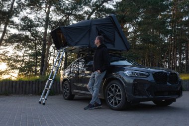 Çatı çadırı ve merdiveni olan modern bir SUV bir ormanda asfalt bir yüzeye park edilmiştir. Uzun çam ağaçlarıyla çevrili, yumuşak ışıkta duran bir adam..