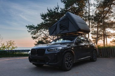 Açık çatı çadırı olan siyah bir SUV, suya yakın bir ormanlık alanda asfalt bir yüzeye park edilmiştir. Uzun ağaçlar ve günbatımı manzarayı çerçeveler..