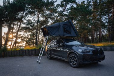 Çatı çadırı ve merdiveni olan modern bir SUV, uzun çam ağaçları ve güneş ışığıyla çevrili bir ormanda asfalt bir yüzeye park edilmiştir..