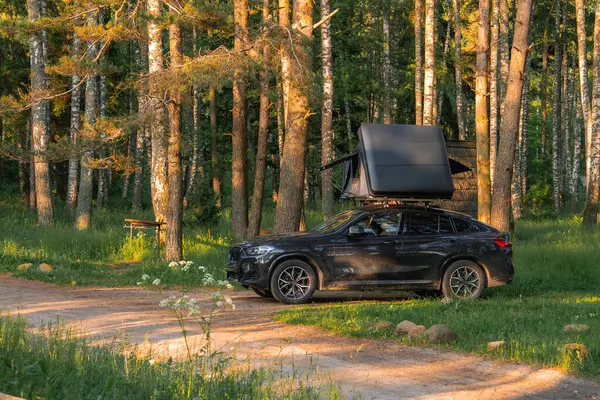 Katlanabilir çatı çadırı olan siyah bir SUV ormandaki kirli bir patikaya park etmiş. Uzun ağaçlar, yeşil yapraklar ve ahşap bir bank huzurlu bir atmosfer yaratır..