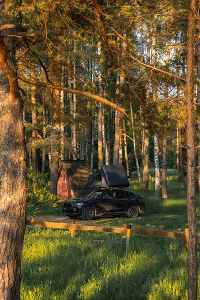 Çadırı kısmen kurulmuş modern siyah bir SUV bir orman açıklığında park halinde. Uzun ağaçlar, ahşap bir kulübe ve sakin sahnenin çerçevesi..