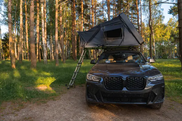 Çatı çadırı ve merdiveni olan modern bir SUV bir orman açıklığında park halinde. Yeşil yapraklı uzun ağaçlar sakin kamp düzenini çevreledi..