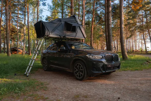Çatısı açık bir çadır ve merdiveni olan bir SUV ormanda park halinde. Güneş ışığı, arka planda görünen ahşap bir kulübeyle uzun ağaçların arasından süzülür..