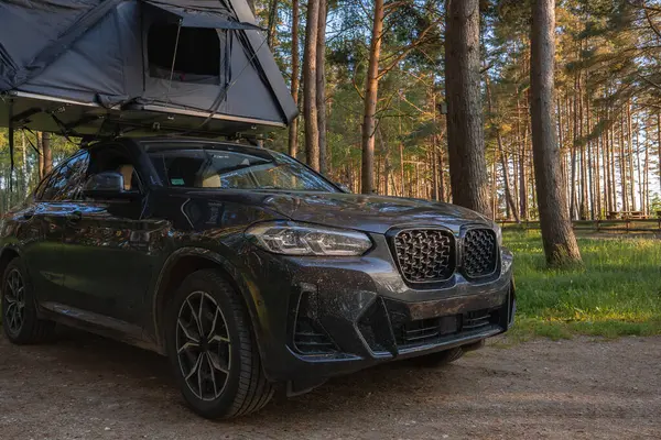 Yarı açık çatısı çadırı olan siyah bir SUV ormanda park halinde. Aracın etrafını saran uzun ağaçlar, gölgelikten süzülen güneş ışığı....