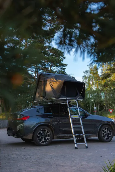 Uzun ağaçlar ve yeşilliklerle çevrili bir ormanda asfalt bir yüzeye park edilmiş katlanabilir çatı çadırı ve metal merdiveni olan modern bir SUV. Bir verim işareti görünür.
