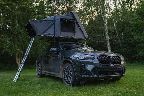 Çimenli bir ormanlık alana kurulmuş katlanabilir çatı çadırı olan modern bir SUV. Uzun yeşil ağaçlarla çevrili bir merdiven yere kadar uzanır..