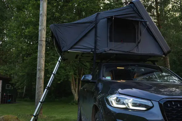 Çatı çadırı ve merdiveni olan modern bir SUV ormanda park halinde. Çevresindeki ağaçlar, bir elektrik direği ve arka planda küçük bir kulübeyle çevrili..