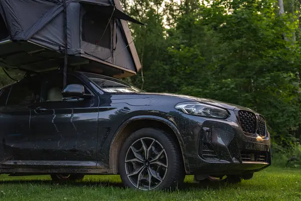 Kısmen açık çatı çadırına sahip modern bir SUV sık bir ormanda çimlerin üzerine park edilmiştir. Araç, doğa ile harmanlanmış, off-road yolculuğundan toprak gösteriyor..