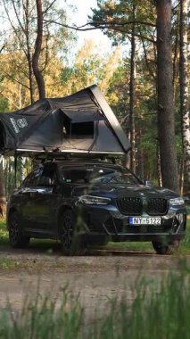 Siyah BMW X4 SUV çatısı çadırı ve merdiveni olan bir orman kampı. Uzun ağaçlar, ahşap bir kulübe ve sıcak güneş ışığı huzurlu bir ortam yaratır..