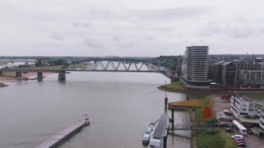 Hollanda, Nijmegen 'in hava manzarası, Waal Nehri, çelik bir köprü, hareketli bir mavna, modern binalar, ve bulutlu gökyüzü altında tarihi mimari.