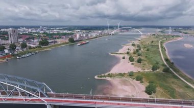 Hollanda, Nijmegen 'in hava görüntüsü Waal Nehri' ni, De Oversteek köprüsünü, hareket eden bir mavnayı, rüzgar türbinlerini, kentsel alanları ve yeşil alanları gösteriyor..