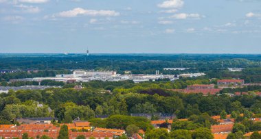 Hollanda, Eindhoven 'ın hava görüntüsü, kırmızı çatılı evleri, endüstriyel binaları, bir iletişim kulesini ve açık bir gökyüzünün altındaki bir ormanı gösteriyor..