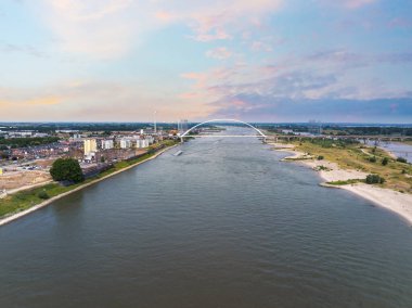 Hollanda, Nijmegen 'in hava manzarası, Waal Nehri üzerindeki De Oversteek köprüsü, kentsel alanlar, yeşil nehir kıyıları ve günbatımında rüzgar türbinleri yer alıyor..