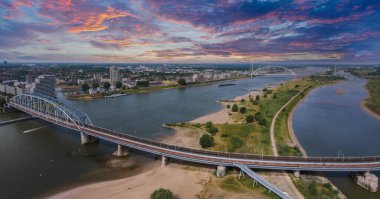 Hollanda, Nijmegen 'in hava manzarası Waalbrug Köprüsü' nü Waal Nehri üzerinde vurguluyor, kumlu nehir kıyıları, yeşil alanlar ve canlı bir günbatımı..
