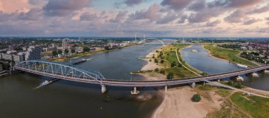 Hollanda, Nijmegen 'in hava görüntüsü Waalbrug köprüsünün Waal Nehri üzerindeki kentsel alanları, yeşil alanları, rüzgar türbinlerini ve parçalı bulutlu gökyüzünü gösteriyor..