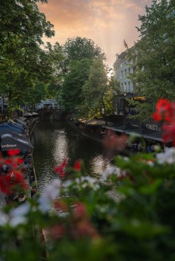 Utrecht, Hollanda 'da tarihi bir köprü, açık kafeler, yeşil ağaçlar, canlı çiçekler ve suya yansıyan altın bir günbatımı bulunan bir kanal..