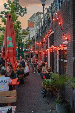 İnsanlar Utrecht, Hollanda 'da dar tuğlalı bir sokak boyunca masalarda, kırmızı ışıklar, şemsiyeler, saksı bitkileri ve Hollanda mimarisi ile yemek yiyorlar..