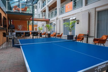 Mavi masa tenisi masası, beyaz duvarlar, cam korkuluklar, turuncu sandalyeler, saksı bitkileri ve ekranda aslan amblemi olan modern kapalı alan..