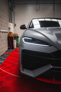 Matte gri Lamborghini Urus kırmızı ve siyah kareli zemine park etmiş. Özellikler açısal tasarım, LED far ve atölye arka plan ayrıntıları.