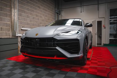 Lamborghini Urus bir garajda mat gümüş kaplama, kırmızı aksanlı, kareli döşeme, beton duvar, beyaz kapı ve görünür elektrik kabloları.
