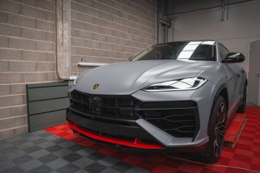 Kırmızı aksanlı gri renkli Lamborghini Urus, beton duvarlar ve borularla çevrili kırmızı ve siyah kareli bir zeminde sergileniyor..