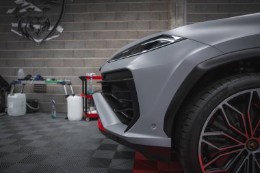 Matte gri Lamborghini Urus ön sağ bölüm keskin farlar, kırmızı aksanlı alaşımlı tekerlek, ve bir garaj arkaplanı ve bir aslan amblemi.