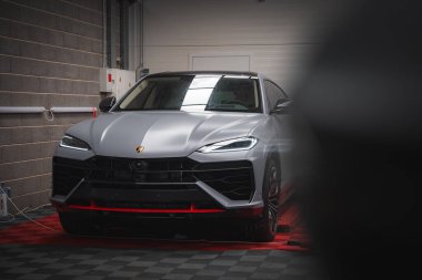 Lamborghini Urus bir garajda mat gümüş kaplama, kırmızı aksanlı, kareli zemin, beton duvar, elektrik panelleri ve tepeden aydınlatma.
