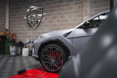Lamborghini Urus 'un ön tarafında gri renkli, siyah ve kırmızı alaşımlı tekerlekler var. Kapalı bir yerde, kırmızı bir paspasın üzerinde gri tuğladan örülü bir duvarı vardı..