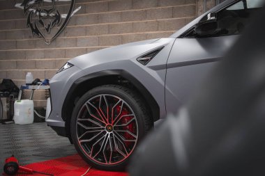 Mat gri renkli Lamborghini Urus 'un yakın görüntüsü, açısal tasarım, kırmızı kalibreli alaşımlı tekerlek, ve boğa amblemli tuğla duvar..