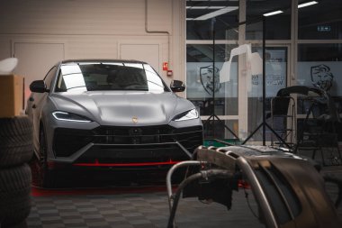 Matte gri Lamborghini Urus. Açısal farları ve bir atölyede amblemi var. Arkaplanda araçlar, lastikler, tampon ve yansımalı cam pencereler yer alıyor.