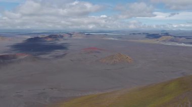 Kırmızımsı bir ovadaki volkanik kraterleri, parçalı bulutlu bir gökyüzünün altında hareket eden gölgelerle çevrili dağlarla süpürüyor..