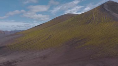 İzlanda 'da volkanik bir arazinin havadan görünüşü, yosun kaplı yamaçlar, kayalık zemin, uzak tepeler ve parçalı bulutlu bir gökyüzü..
