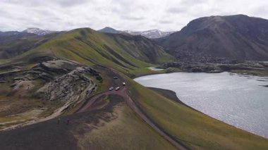 Yeşil yosunlar, volkanik kayalar, dingin bir göl ve karla kaplı zirvelerle çevrili bir tepenin üstündeki, dolambaçlı toprak bir yolda uçan araçların görüntüsü..