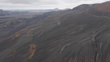 İzlanda 'daki volkanik bir dağın üzerinden uçan hava aracı kül kaplı yamaçlar, turuncu bitki örtüsü ve bulutlu bir gökyüzünün altında kar tepeleri..