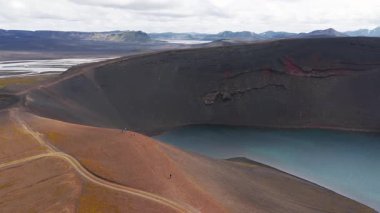 Mavi suyu, kırmızı çizgili duvarları ve toprak yolu olan volkanik bir krater gölü. Kar kaplı dağlar uzaklardan görülebilir..