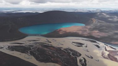 Karanlık volkanik arazilerle çevrili canlı bir mavi krater gölü, nehirler, engebeli yamaçlar ve bulutlu bir gökyüzünün altındaki uzak dağlar..