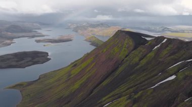İzlanda 'da uçsuz bucaksız bir göl manzarası engebeli, çok renkli dağlar, yeşil yosun, volkanik kaya ve gökyüzünde soluk bir gökkuşağı ile çevrili..