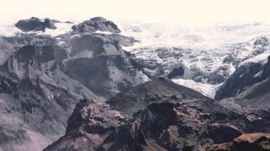 İzlanda 'daki karanlık volkanik dağlar, buzlu genişlikler, kayalık arazi ve yeşil bitki örtüleri arasında uzanan bir buzulun havadan görünüşü..
