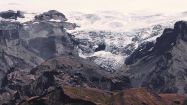 İzlanda 'daki karanlık volkanik kayalıkların arasına yerleşmiş bir buzulun hava manzarası, yeşil bitki örtüleri ve manzarayı gösteren yavaş bir tava..