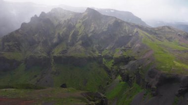 İzlanda 'nın engebeli arazisindeki sisli ve karlı zirvelerle çevrili yemyeşil bir vadiyi yavaş yavaş süpüren bir hava manzarası..