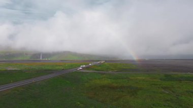 Düz bir yol, yemyeşil İzlanda manzarasının içinden geçer. Uzakta çağlayan şelaleler ve hareket halindeki arabalarla gökkuşağı kemerleri..