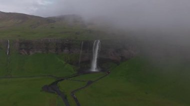 Seljalandsfoss şelalesi İzlanda 'da yemyeşil bir araziyle çevrili yüksek bir uçurumdan dökülüyor. Yollar ve ziyaretçiler görünür, olay yerine hareket katıyorlar..
