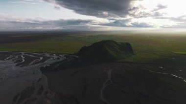 İzlanda 'nın düz ovalarından yükselen yeşil bir tepenin havadan görünüşü, karanlık nehir yatakları ve bulutların ve güneş ışığı parçalarının olduğu dramatik bir gökyüzü..