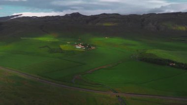 Engebeli dağları olan yemyeşil bir vadinin havadan görünüşü, arka planda bir buzul, tarlaları ve toprak yolları olan bir kırsal yerleşim yeri. Yavaş süpürme hareketi.
