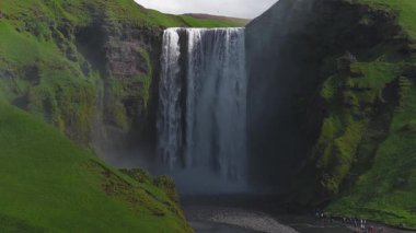 İzlanda 'daki Skogafoss şelalesinin hava manzarası, aşağıdaki sisli havuza dökülen su, yemyeşil tepeler ve engebeli araziyle çevrili..