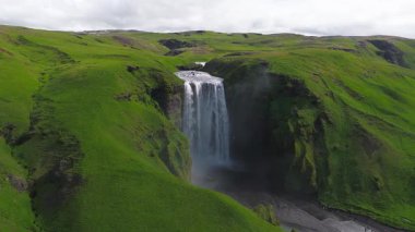 İzlanda 'daki Skogafoss şelalesinin hava manzarası sisli bir havuza dökülüyor. Patikalar ve yollar görülebilir, engebeli tepeler ve uzak dağlarla.