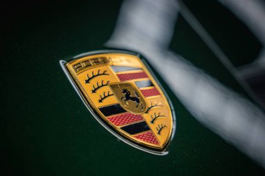 Işıl ışıl yeşil bir arabanın kaputundaki Porsche ambleminin detaylı bir görüntüsü, iyi aydınlatılmış kapalı bir garajdaki tepedeki ışıkların yansımaları..