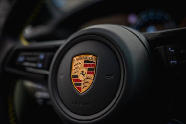 İkonik armayı, ayrıntılı dikişleri ve aydınlatma göstergeleriyle bulanık gösterge paneli elemanlarını içeren bir Porsche direksiyonunun yakın görüntüsü.